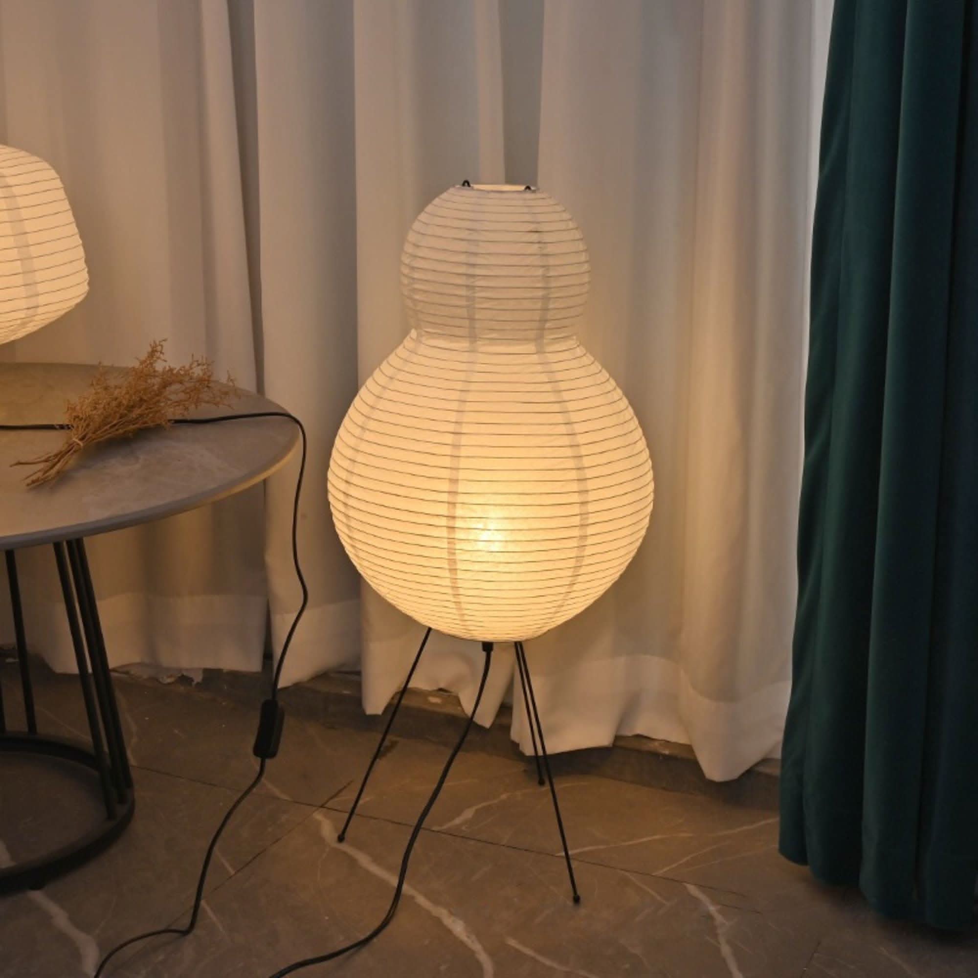 White paper floor lamp with pear shape, on black tripod legs, beside a table.